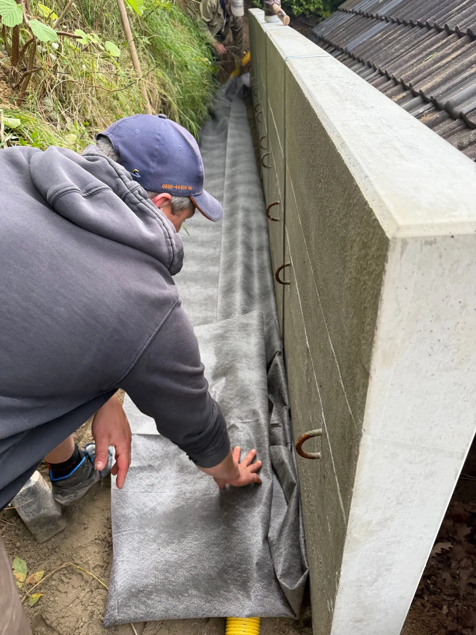 Mitarbeiter legt Filtervlies hinter der Stützwand aus, bevor das Drainagerohr eingebettet wird.