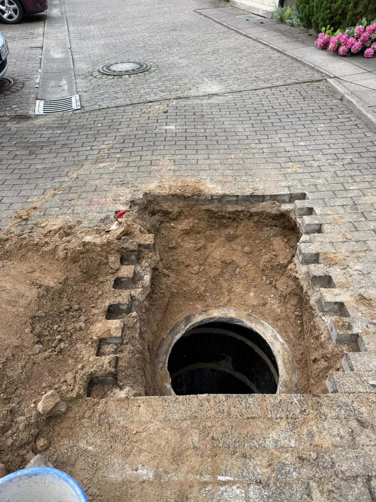 In einer aufgenommenen Pflasterflaeche freigelegter Kanalanschluss mit offener Rohrmuffe, die Pflastersteine sind stufig und sauber ausgebaut.