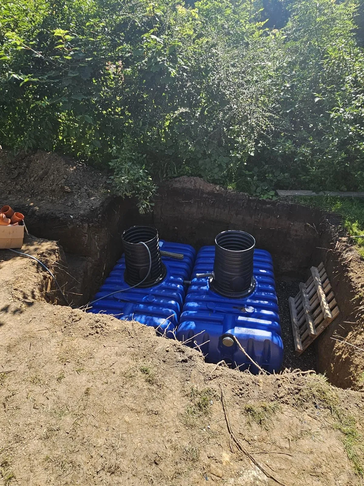Blick von oben in die Baugrube auf zwei nebeneinander gesetzte blaue Regenwassertanks mit schwarzen Schachtrohren, gebettet auf Kies.