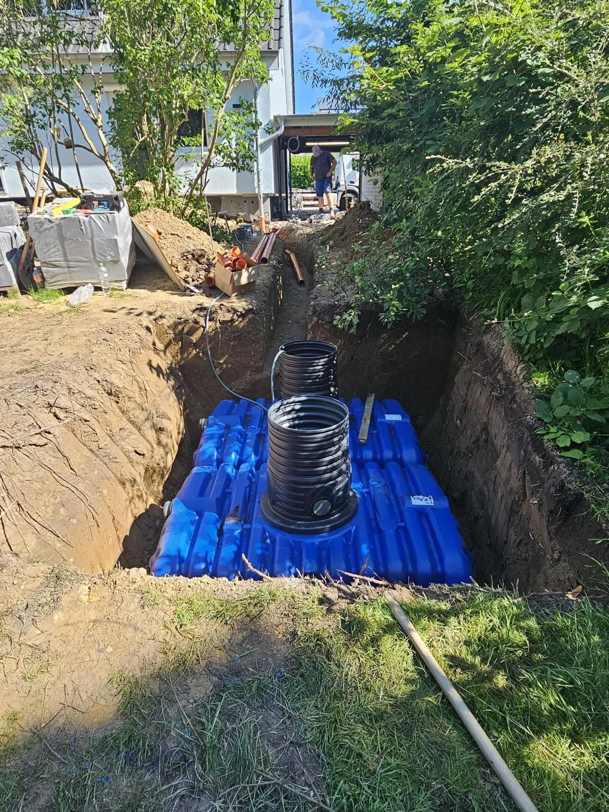 Zwei blaue Versickerungstanks liegen in der ausgehobenen Grube neben dem Wohnhaus, darauf zwei schwarze Schachtaufsaetze zur Kontrolle.