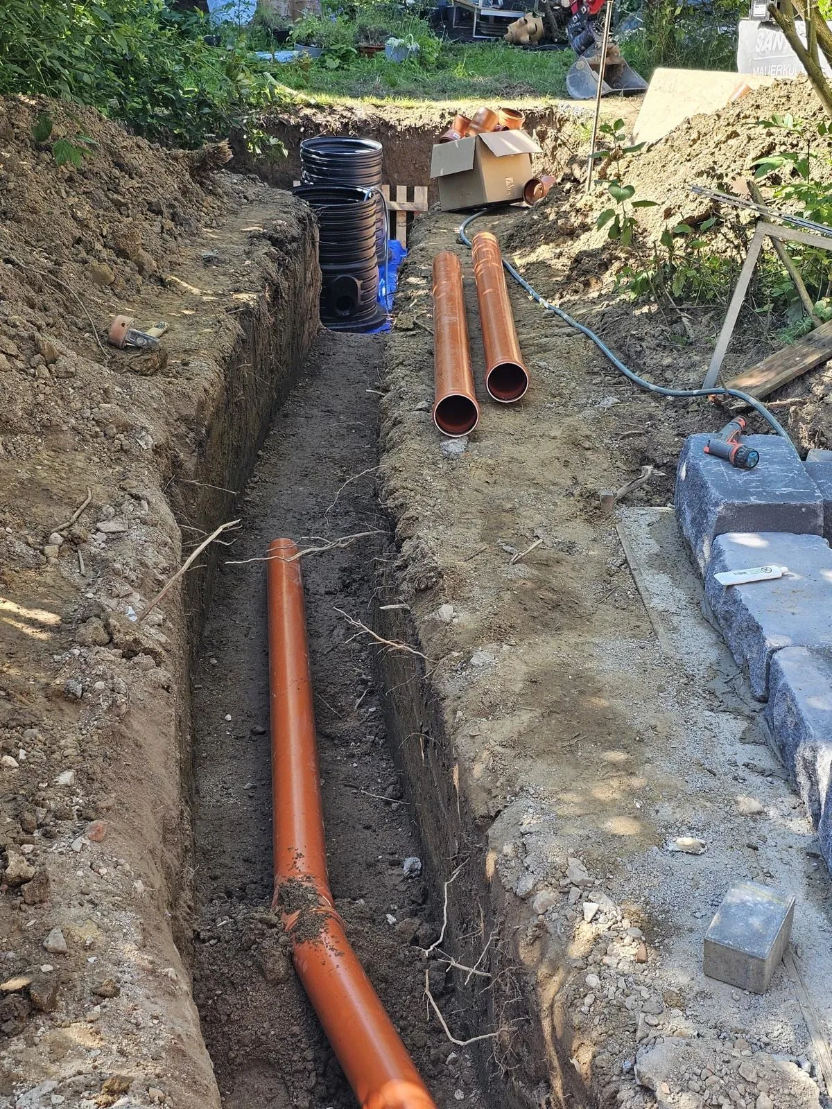 Ausgehobener Rohrgraben mit bereits verlegter orangefarbener Leitung, am Grabenrand warten Schachtringe und weitere Rohre auf den Einbau.