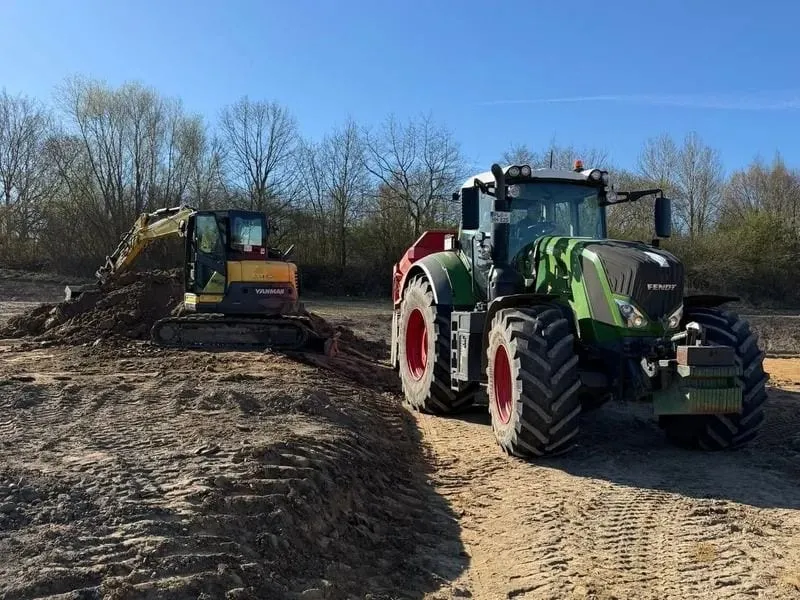 Bagger modelliert eine Erdfläche, daneben wartet ein Traktor mit Muldenkipper.