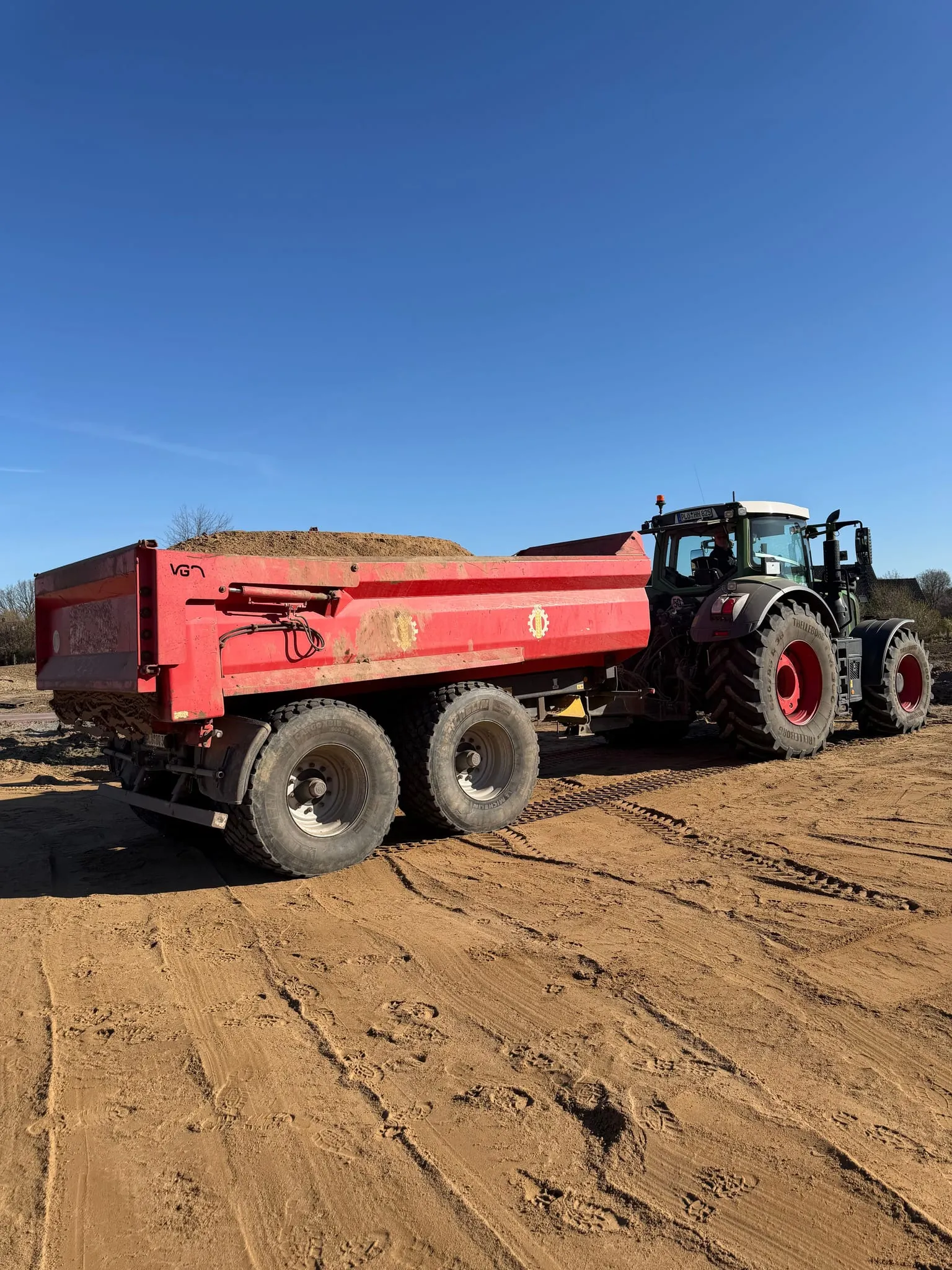 Traktor mit rotem Muldenkipper auf einer planierten Sandfläche.