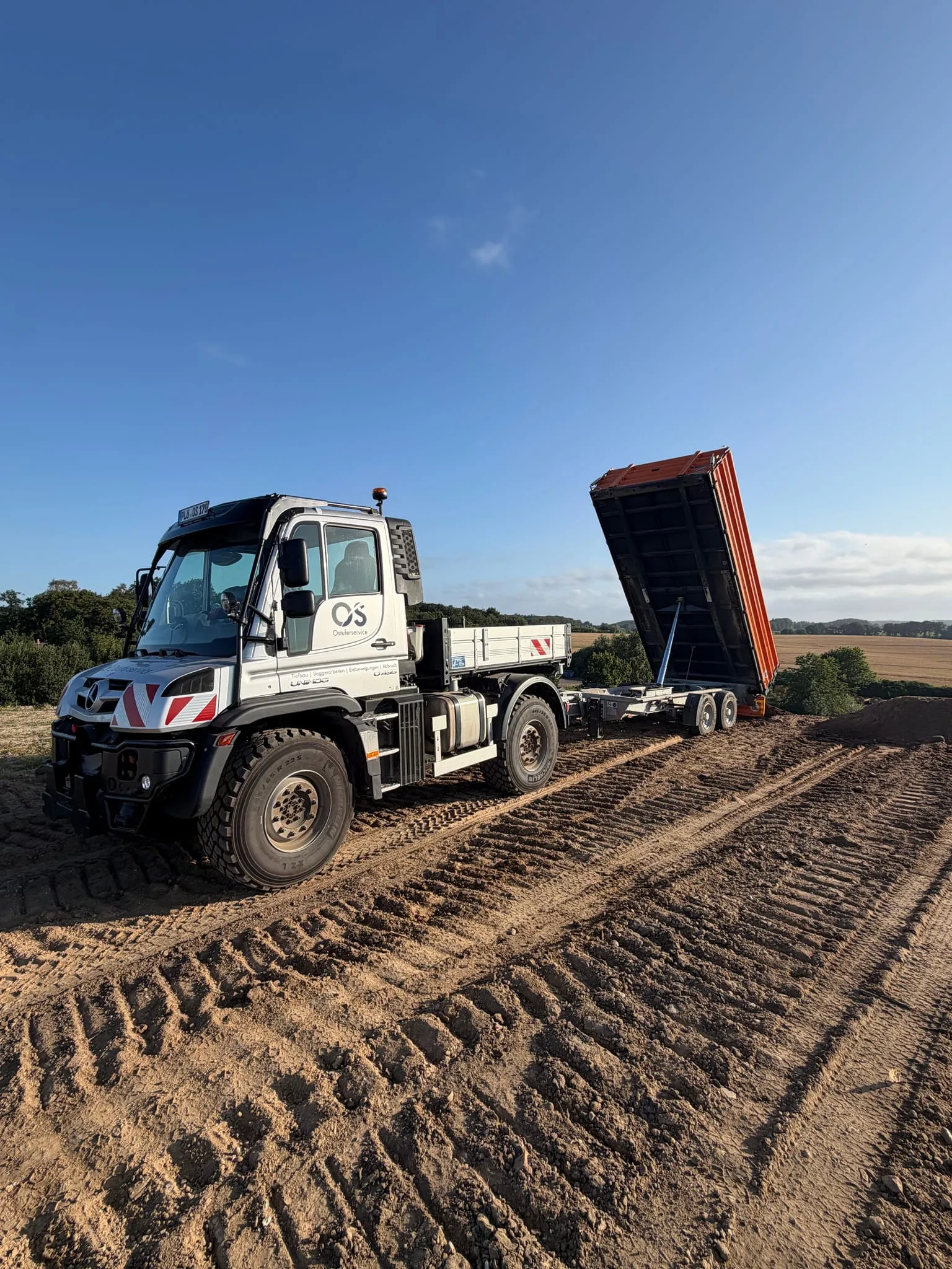 Unimog mit Kippanhänger auf einer frisch bearbeiteten Fläche unter blauem Himmel.