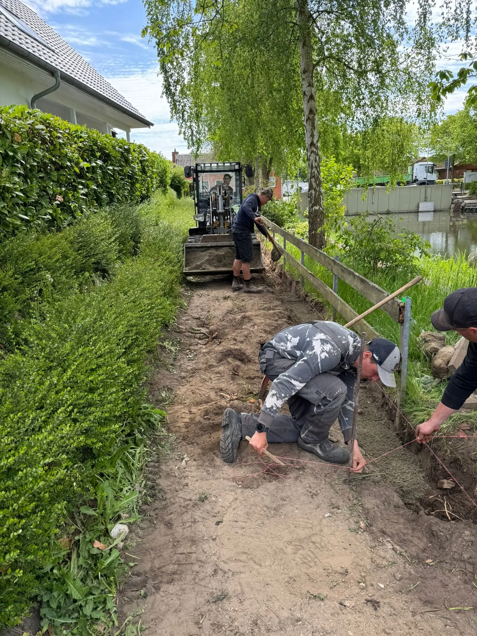 Drei Mitarbeiter bauen den Weg ein: Einer verteilt Sand aus der Radladerschaufel, zwei richten am Boden die Schnur fuer die Kante aus, im Hintergrund der Teich.