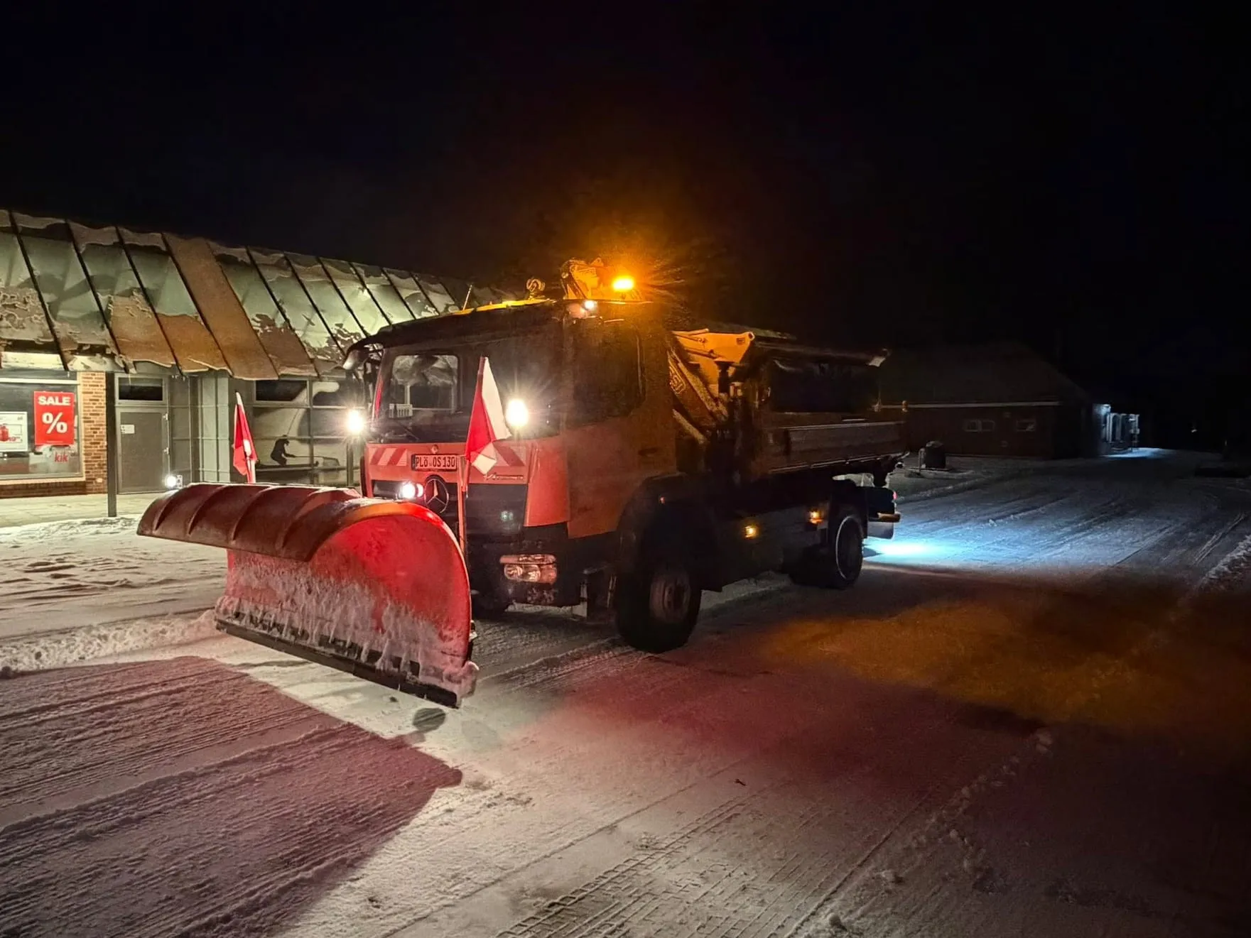 Räumfahrzeug mit beleuchtetem Schneepflug schiebt nachts den Schnee von einem Parkplatz