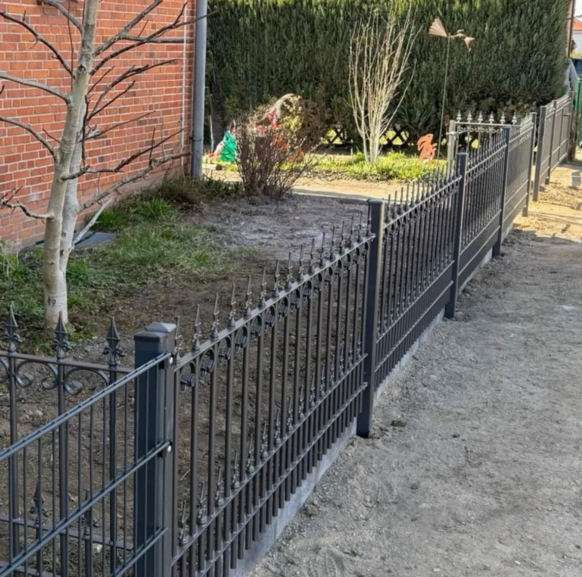 Frisch gesetzter anthrazitfarbener Schmuckzaun aus Metall mit Zierspitzen entlang einer Grundstücksgrenze, die Pfosten stehen in frischem Beton.